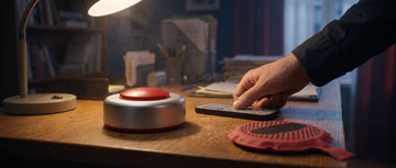 A remote controlled fart machine, its remote and a red whoopee cushion on a desk